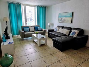 A living room with a sofa, a coffee table, and a TV. There is a window with curtains, a lamp, and a painting on the wall. - Vive la experciencia frente al mar (Hollywood)
