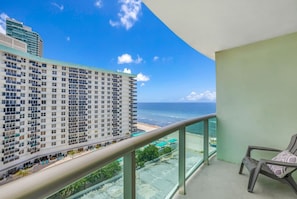A balcony with a view of a beach and ocean, a chair, and a building with multiple floors. - Vive la experciencia frente al mar (Hollywood)