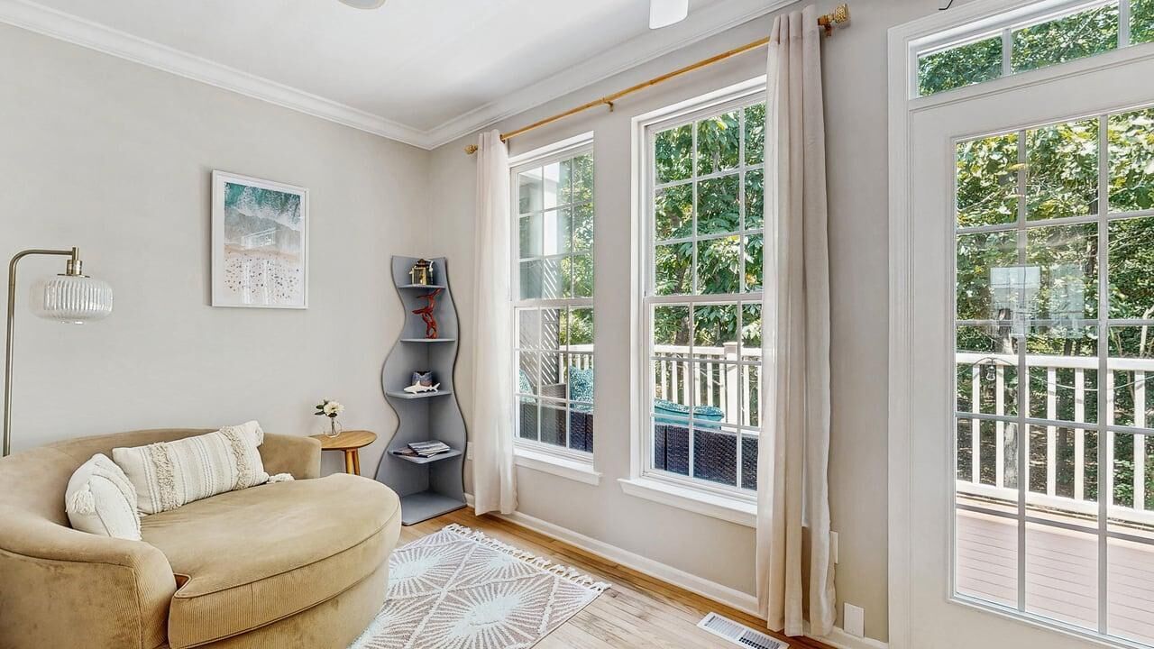 Une pièce avec un canapé beige, une petite table d’appoint, un lampadaire et une grande fenêtre donnant sur un paysage verdoyant.