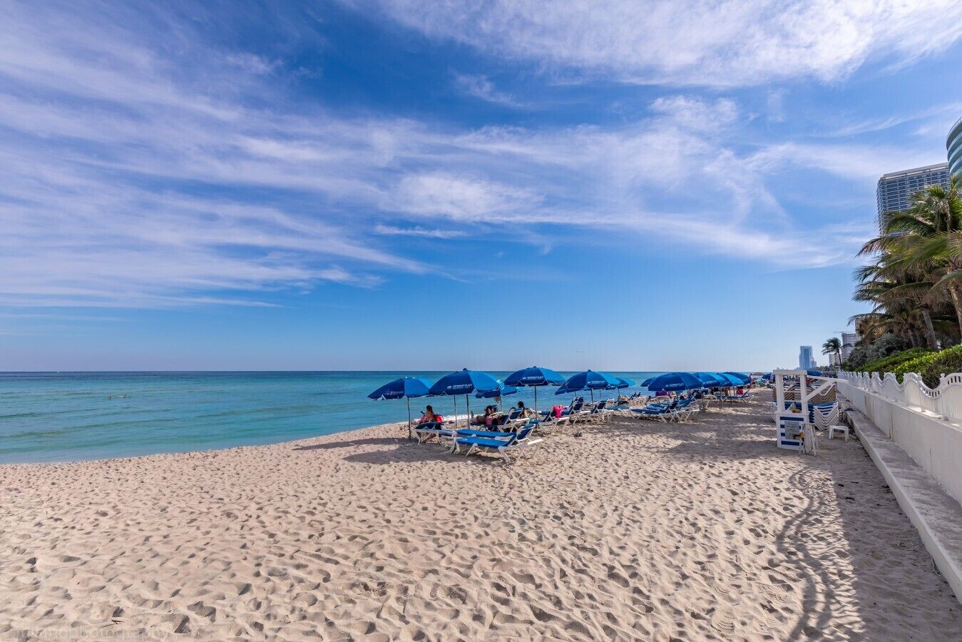 Uma praia com espreguiçadeiras e guarda-sóis, céu claro e mar calmo.