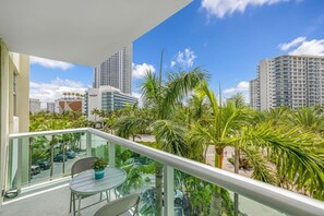 A balcony with a glass railing, a table, and chairs, overlooking a parking area and palm trees with tall buildings in the background. - Hollywood Escape |Piscina| Playa | Vistas | Balcón (Hollywood)