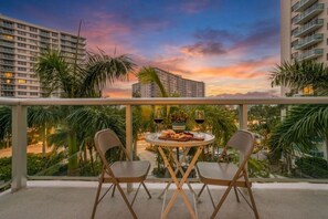 Un balcon avec deux chaises et une table dressée pour un repas, offrant une vue sur le paysage urbain au coucher du soleil.