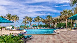 Zona de piscina en un resort, con sillas de descanso, sombrillas y palmeras.