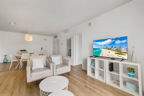 Un salon moderne avec un téléviseur à écran plat, des meubles blancs et une décoration murale sur le thème de la plage.