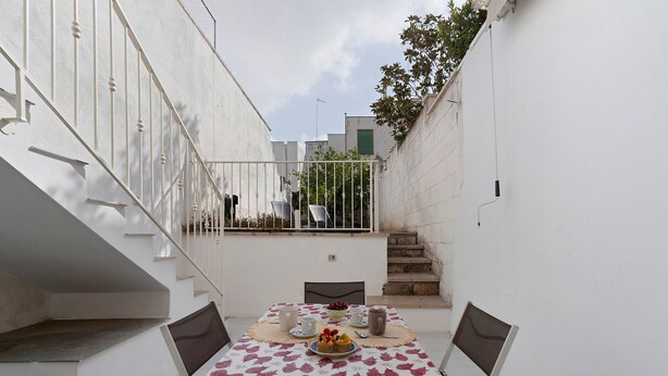 Een smal buitenterras met een tafel voor twee personen, een witte balustrade en een trap naar een balkon.