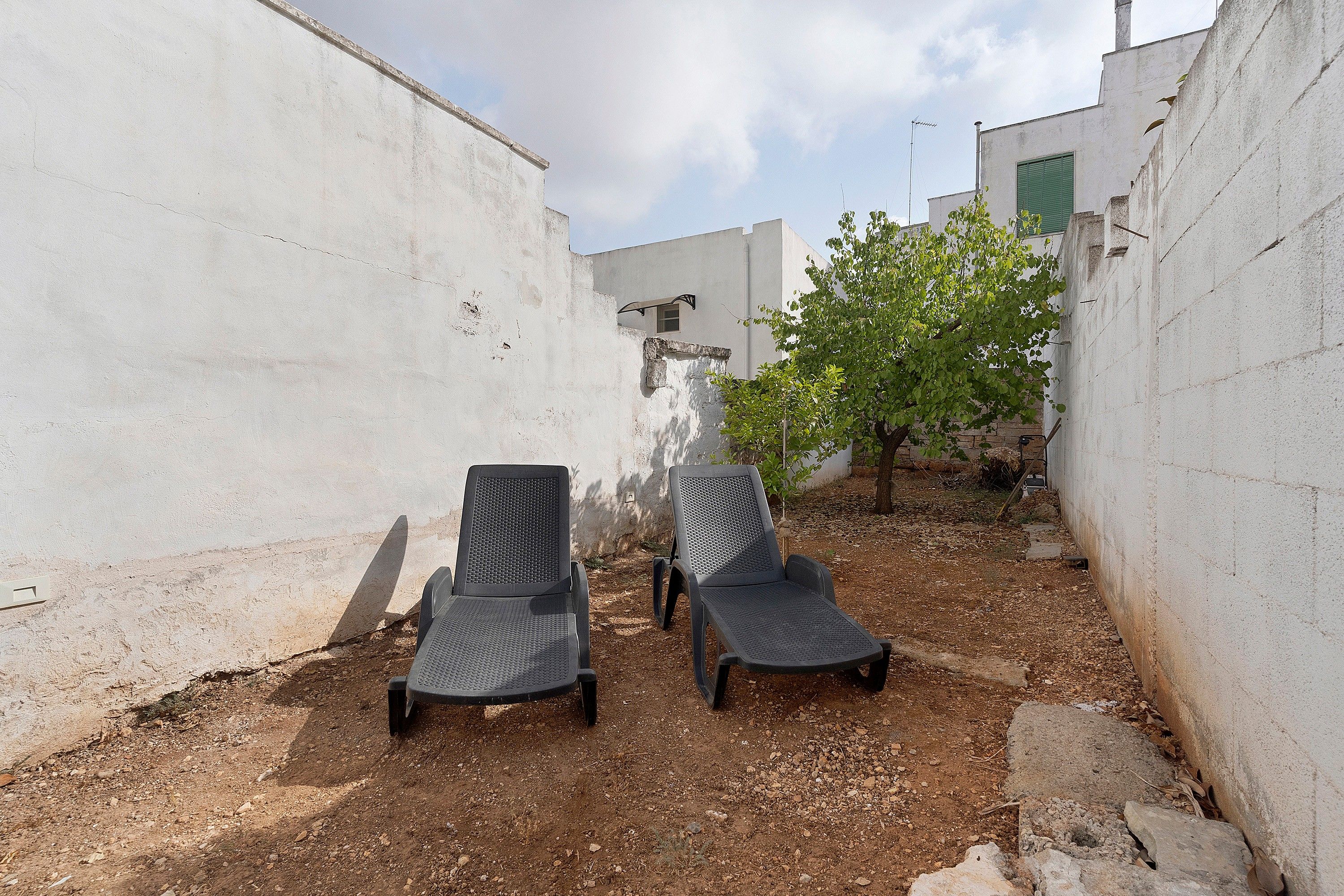 Dos sillas de exterior en un patio estrecho con paredes blancas y un pequeño árbol.