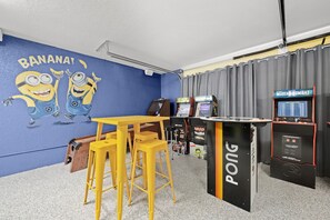 Habitación con pared azul adornada con un mural de Minions, una mesa amarilla con taburetes y máquinas de videojuegos, incluyendo un juego de Pong.