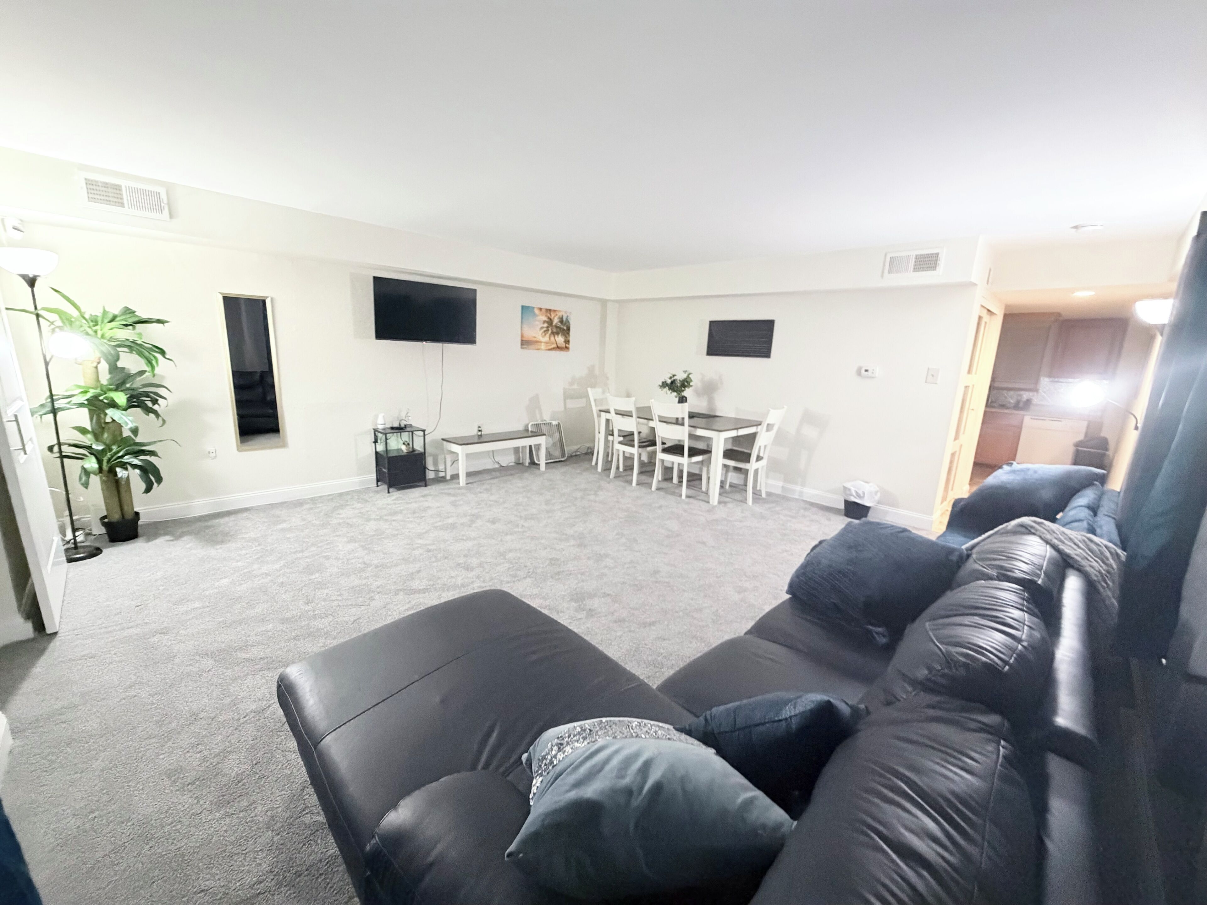 Ein Wohnzimmer mit einer schwarzen Ledersofa, einem Esstisch mit Stühlen, einem an der Wand montierten Fernseher und einer Topfpflanze.