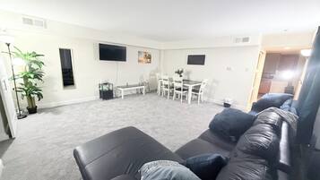 Ein Wohnzimmer mit einer schwarzen Ledersofa, einem Esstisch mit Stühlen, einem an der Wand montierten Fernseher und einer Topfpflanze.