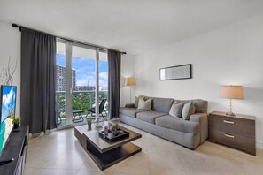A modern living room with a grey sofa, a coffee table, a TV, and a balcony with a view of buildings and greenery. - Hollywood Oasis Familiar |Playa | Piscina | Balcón (Hollywood)