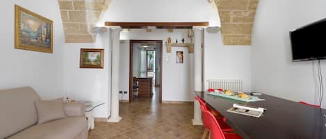A room with a stone archway, a sofa, a dining table with red chairs, and a television mounted on the wall.