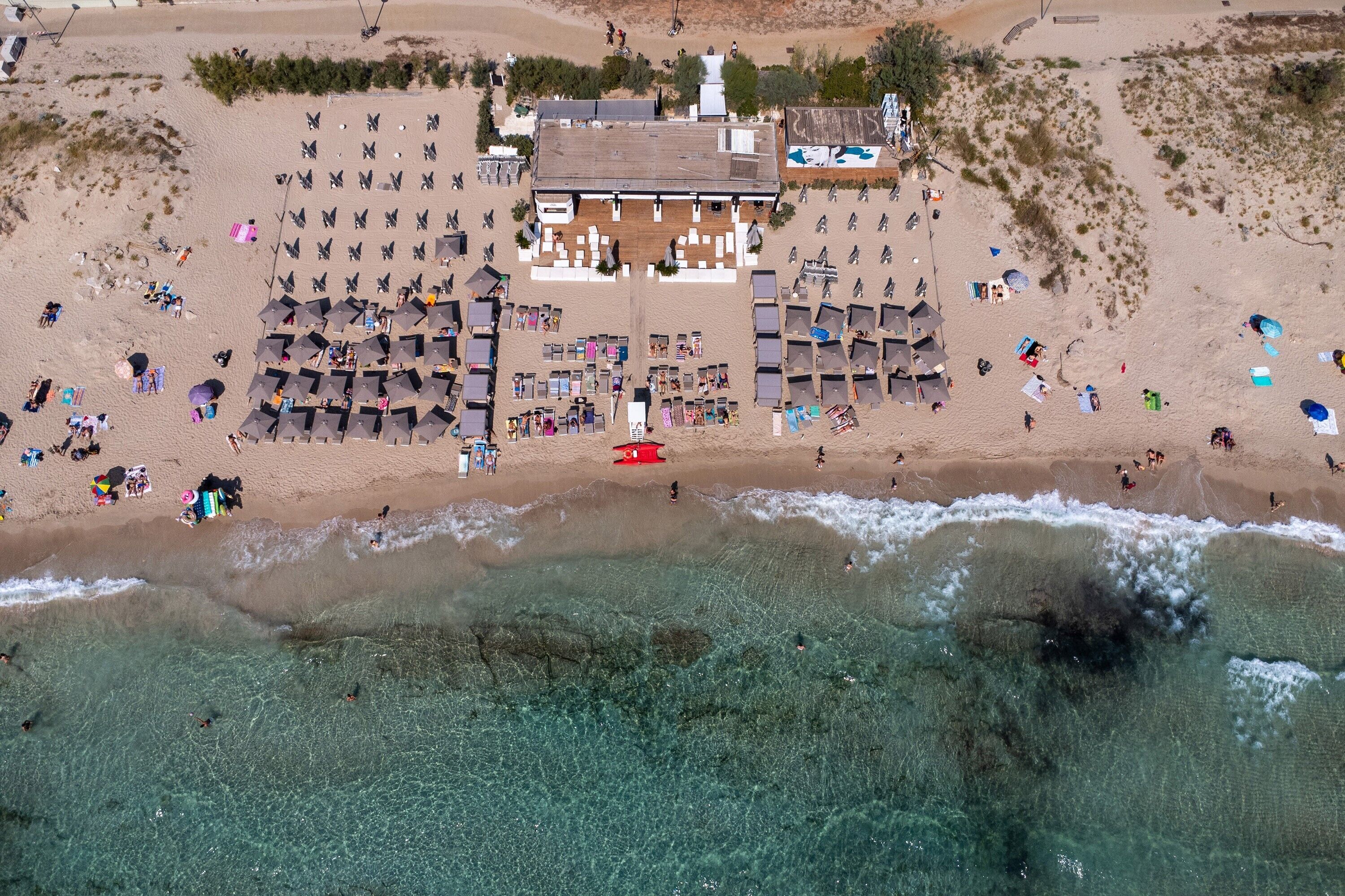 An aerial view of a beach resort with multiple sun loungers, a central building, and clear turquoise waters.