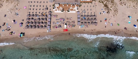 An aerial view of a beach resort with multiple sun loungers, a central building, and clear turquoise waters.