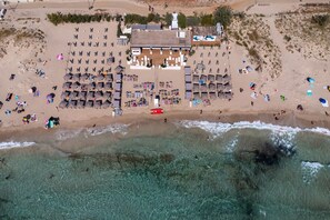Vista aérea de un complejo turístico en la playa, con múltiples tumbonas, un edificio central y aguas turquesas cristalinas.