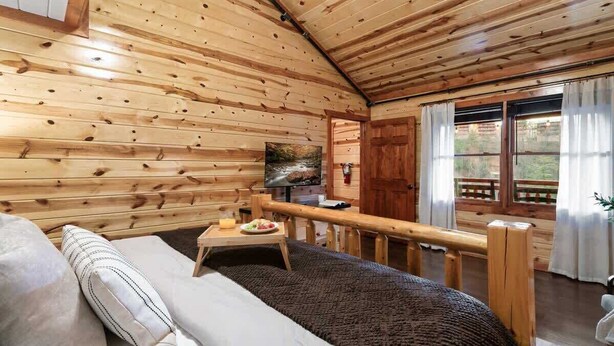 Interior de una cabaña de madera con un sofá, una mesa de comedor con un plato de comida, un televisor y una ventana con cortinas.