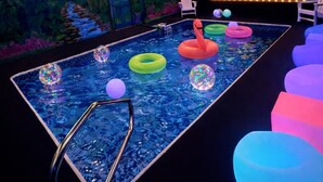 Una piscina con anillos de colores flotando, un letrero de neón que dice 'POOL CINEMA' y un mural de un atardecer.