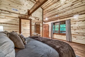 Ein Schlafzimmer in einer Blockhütte mit einem großen Bett, einer Holzbalkendecke und einem Fenster mit Vorhängen.
