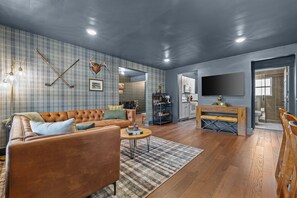 A modern living room with a brown leather sofa, a coffee table, and a television. - Private Mini Golf Course, Main House + Clubhouse, HotTub, Games, Walk to Bars (Nashville)