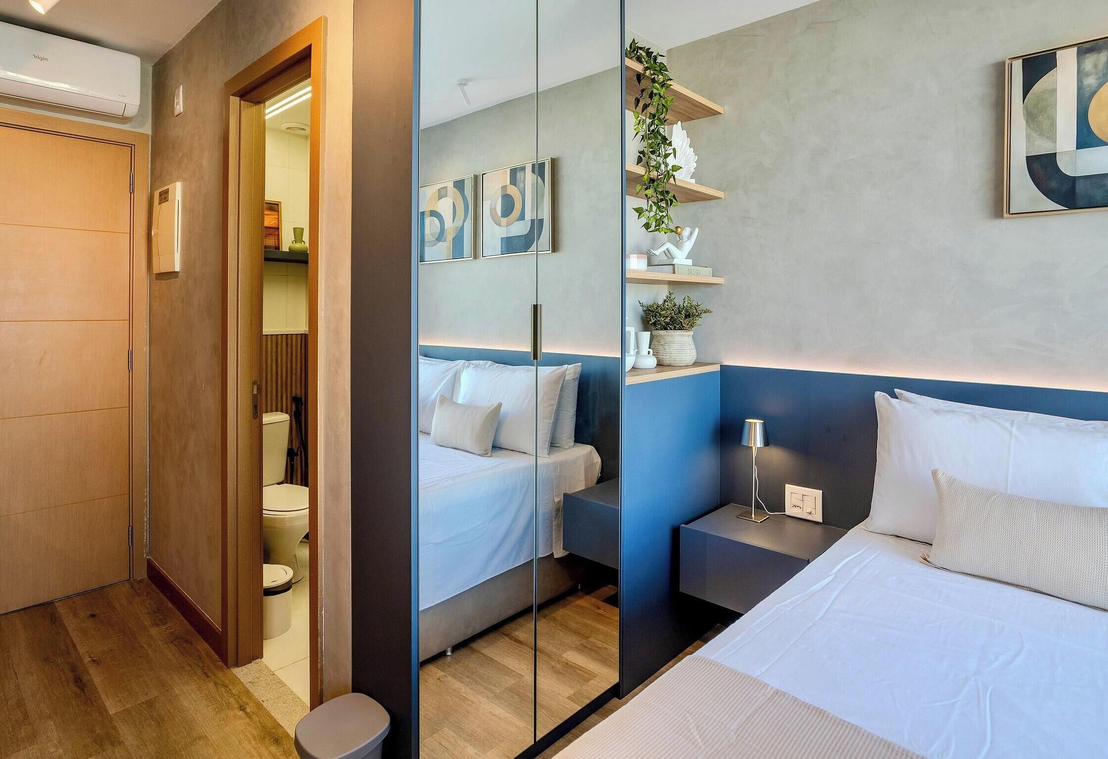Una habitación de hotel moderna con un espejo grande, una cama, mesitas de noche y un baño pequeño.