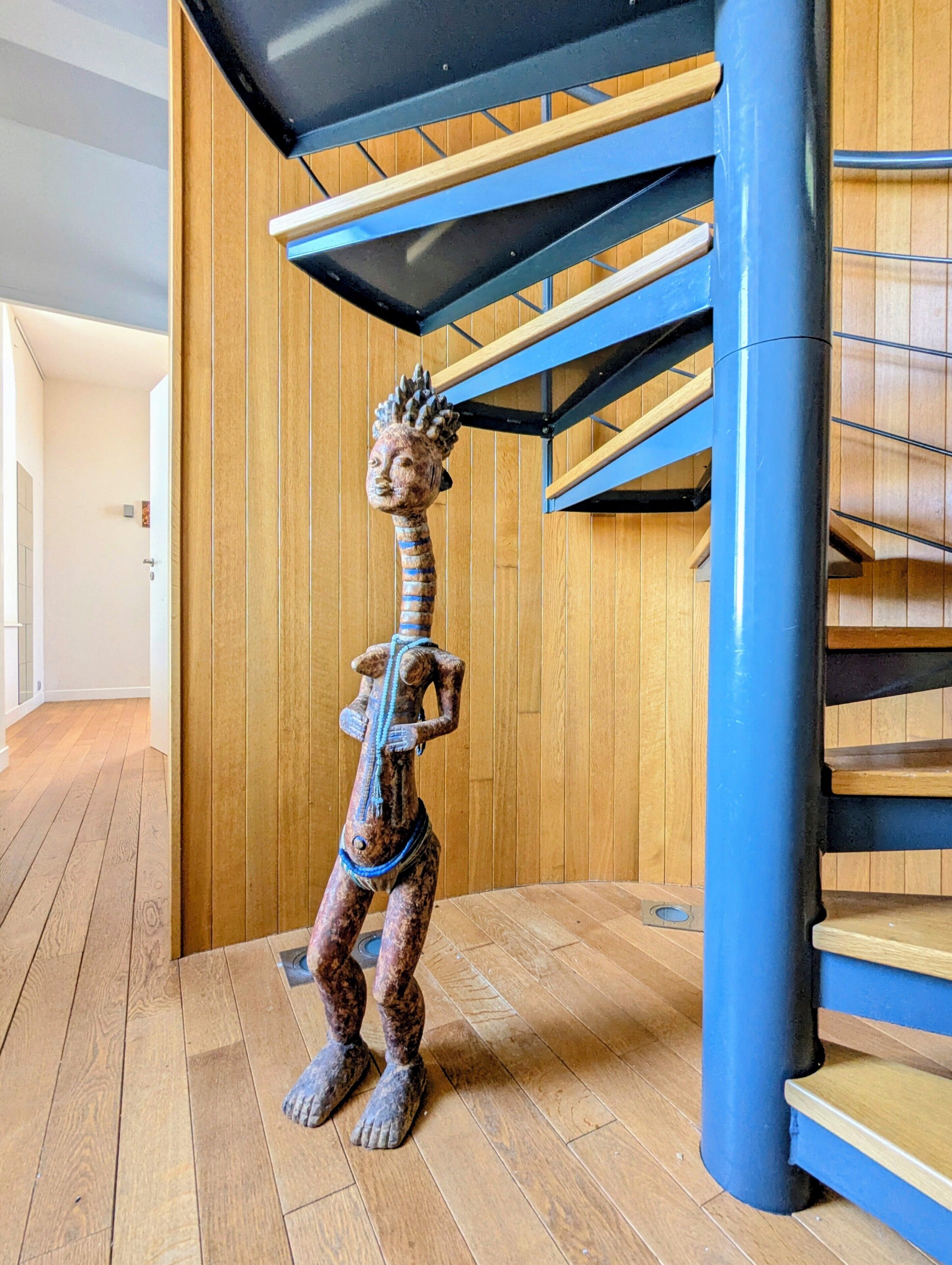 Eine Skulptur einer Menschengestalt mit Kopfschmuck, vor einer Holzwand und einer blauen Treppe platziert.