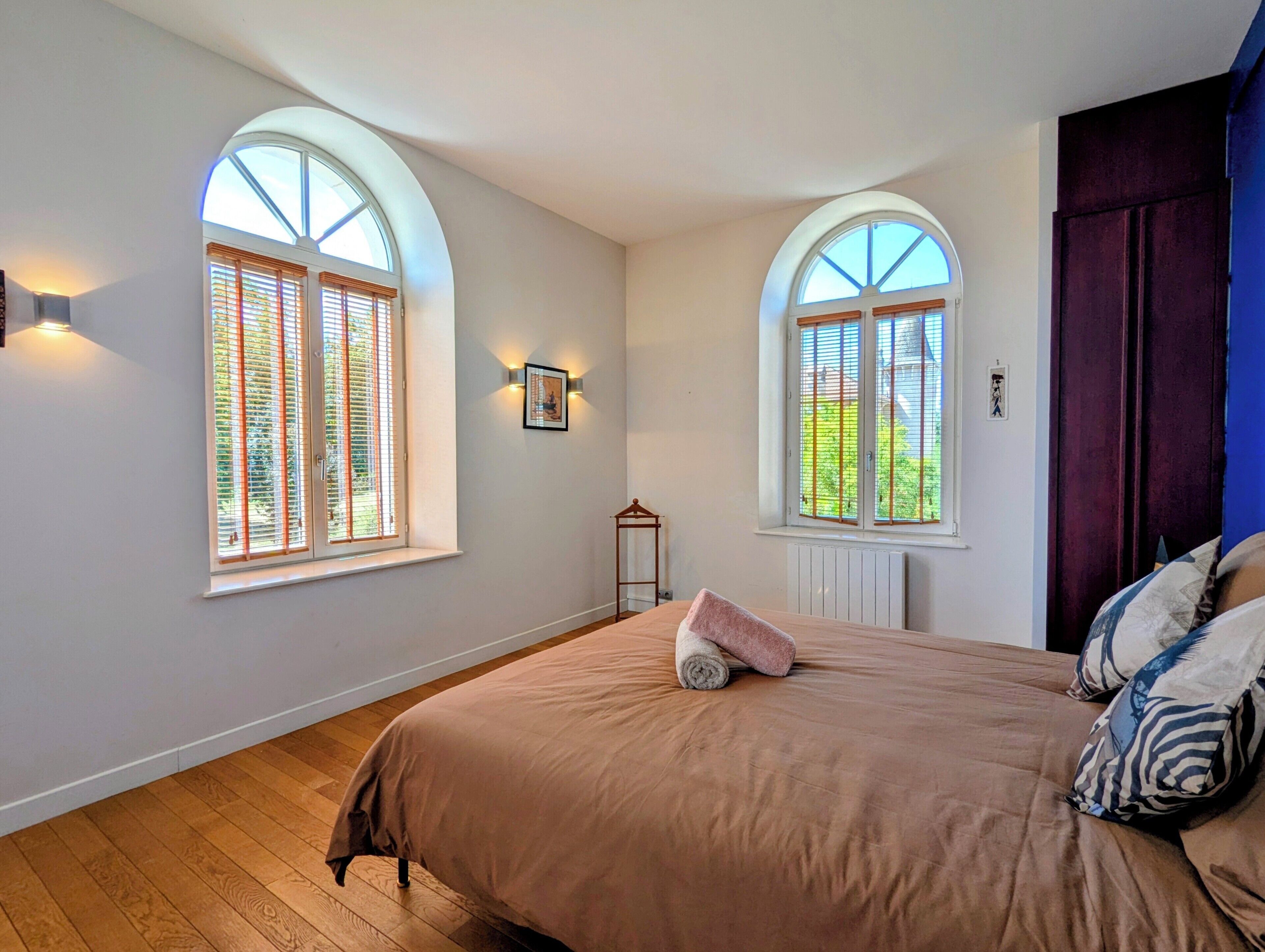 Un dormitorio con una cama grande, dos ventanas con persianas, un radiador y un armario de color oscuro.