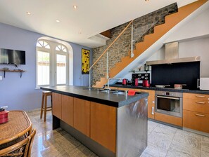 A modern kitchen with a central island, a stone staircase, and a dining area with a wicker table. - Villa Teranga - Terre d'Accueil - Château Colin (Belleau)