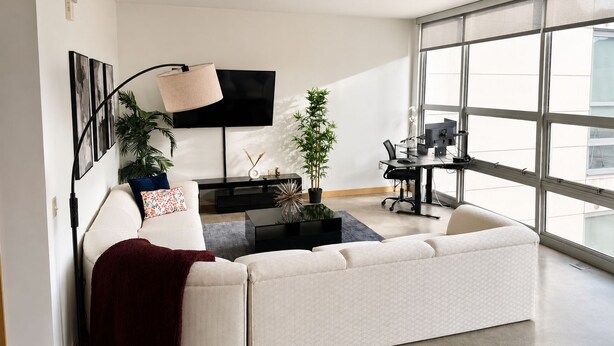 A modern living room with a large window, a flat-screen TV, a sofa, a coffee table, and a desk with a computer. - Modern Downtown Detroit Loft with Skyline views (Detroit)