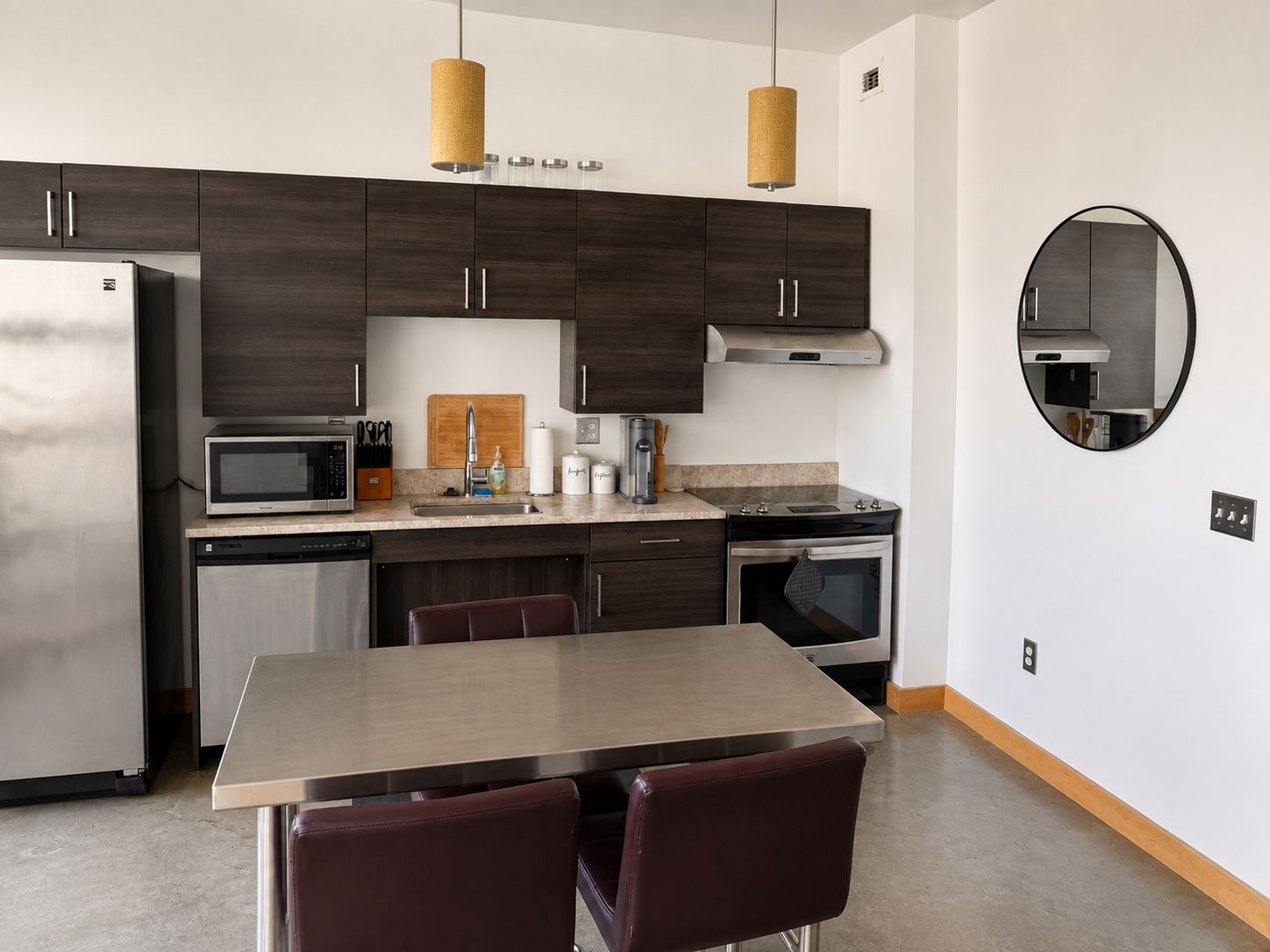 A modern kitchen with dark wood cabinets, stainless steel appliances, a round mirror, and a dining area with a wooden table and leather chairs.