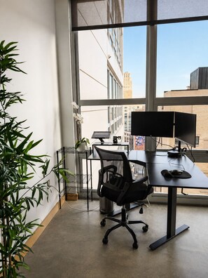 A modern office space with a desk, chair, computer, and a large window offering a view of buildings.