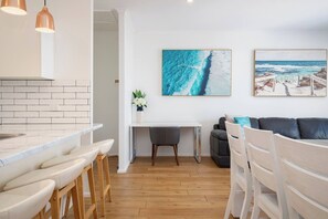A modern kitchen with white brick backsplash, wooden bar stools, a dining table with chairs, a white vase with flowers, and two paintings on the wall. - Sandy Shores A Spacious Newcastle Beach Escape (Newcastle)