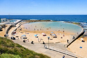 A coastal beach with a curved pool, sandy areas, and a promenade. - Sandy Shores A Spacious Newcastle Beach Escape (Newcastle)