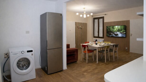 Un espacio habitable compacto con lavadora, refrigerador, comedor con mesa y sillas, televisor montado en la pared y piso de madera.