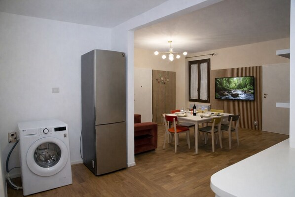 A compact living space with a washing machine, a refrigerator, a dining area with a table and chairs, a television mounted on the wall, and a wooden floor.