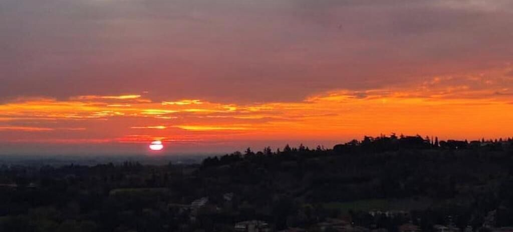Een zonsondergang boven een glooiend landschap, met silhouetten van bomen en gebouwen.