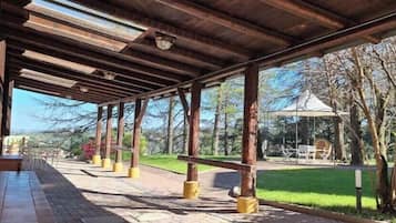 An outdoor covered area with wooden beams and columns, a pathway, and a view of a park with trees and a gazebo.