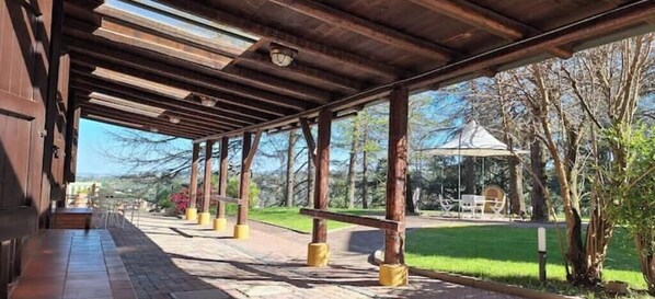 Un espace extérieur abrité, doté de poutres et de colonnes en bois, un sentier et une vue sur un parc avec des arbres et un kiosque.