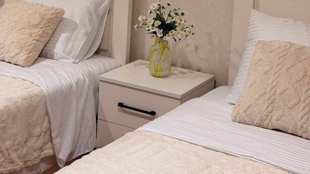 A bedroom with two beds, each with a white headboard and a beige quilted blanket. There is a nightstand with a vase of white flowers between the beds. - Horizont Boutique Hotel (Telavi)