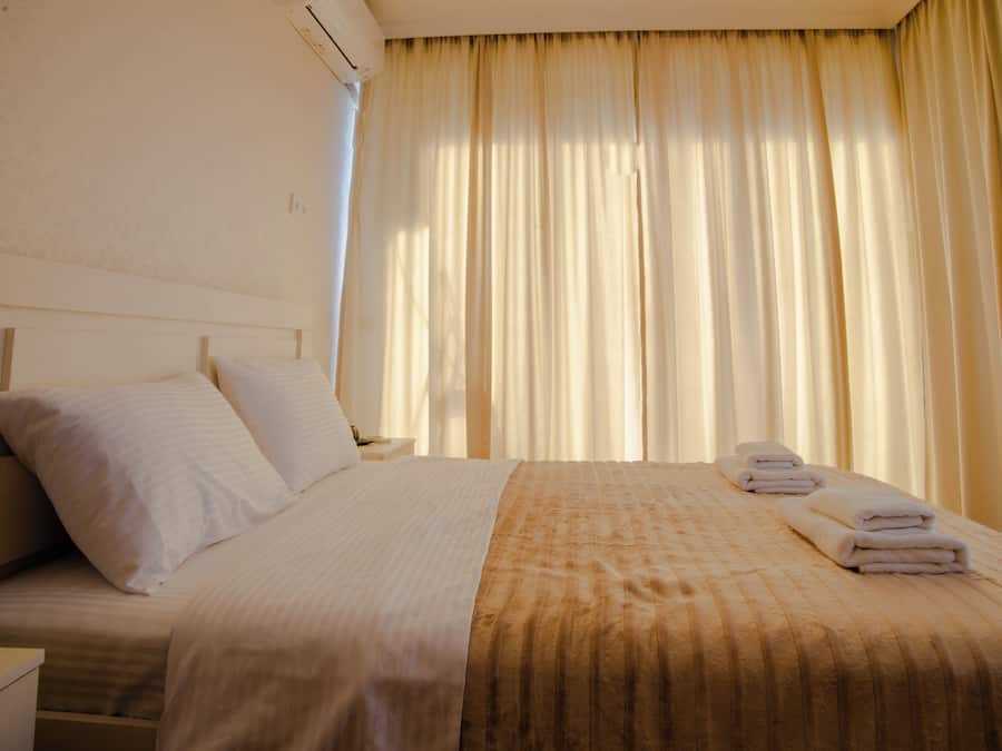 A hotel room with a bed, pillows, a nightstand, a lamp, and a window with curtains.