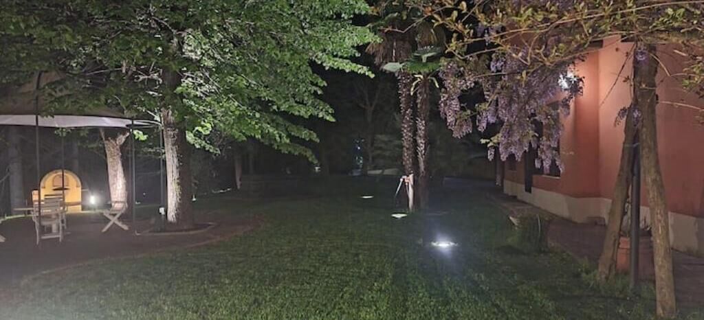 Área externa iluminada à noite, com gramado, árvores e uma estrutura com luzes.