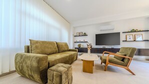 Ein modernes Wohnzimmer mit einer Sofagarnitur, einem Couchtisch und einem Sessel. An der Wand ist ein Flachbildfernseher angebracht und ein kleiner Tisch mit dekorativen Gegenständen.