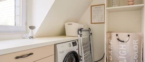 Un espace dédié à la lessive, comprenant un lave-linge, un étendoir, un panier et une étagère avec un sac à linge et un tableau illustré indiquant les instructions de lavage.