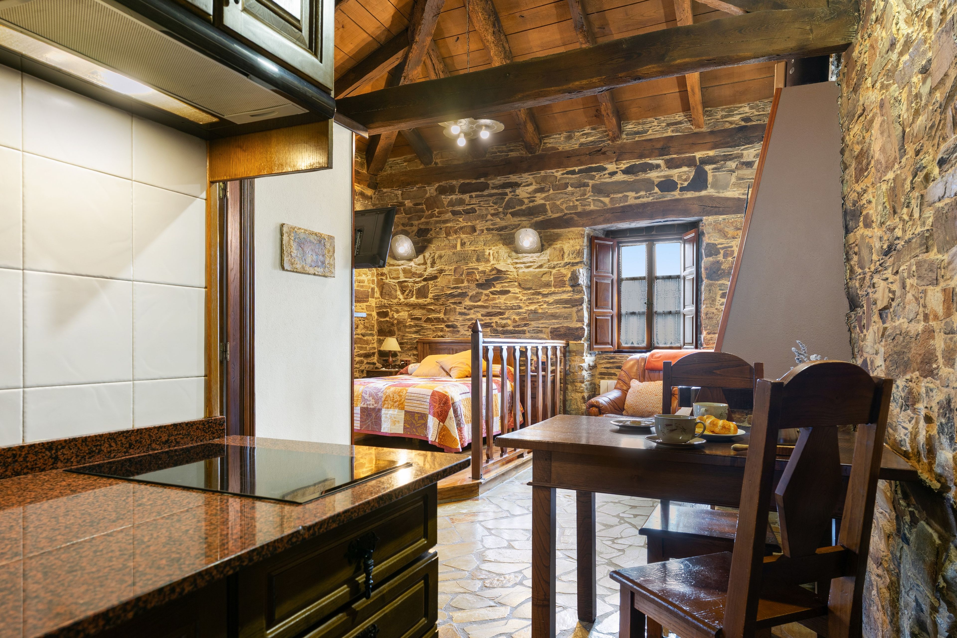 Un interior rústico con pared de piedra, vigas de madera, suelo de baldosas, cama con colcha colorida, mesa de madera con una taza y un pastel, y ventana con vistas.
