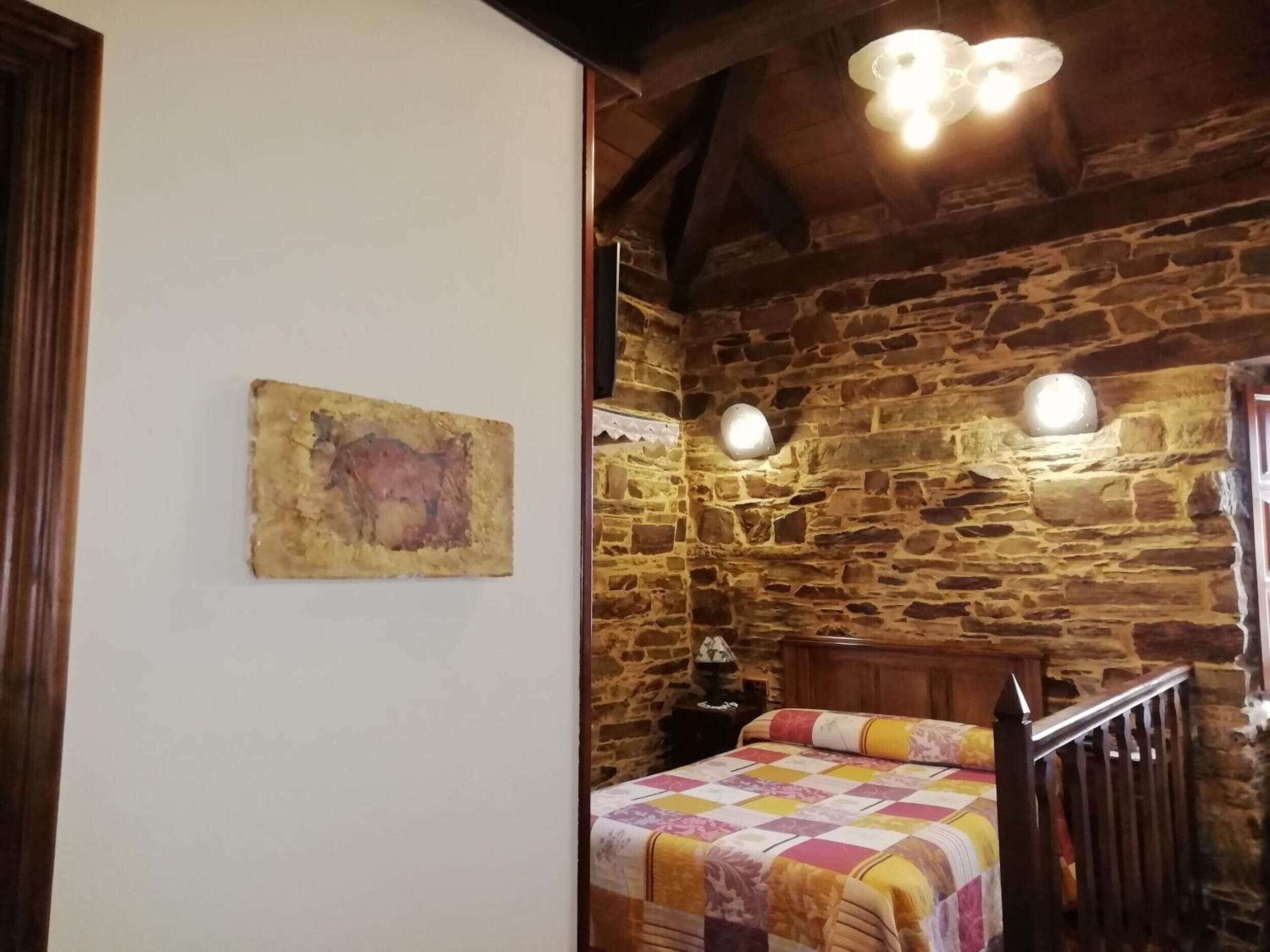 A rustic room with a stone wall, a wooden bed with a colorful quilt, a wall-mounted light fixture, and a painting.