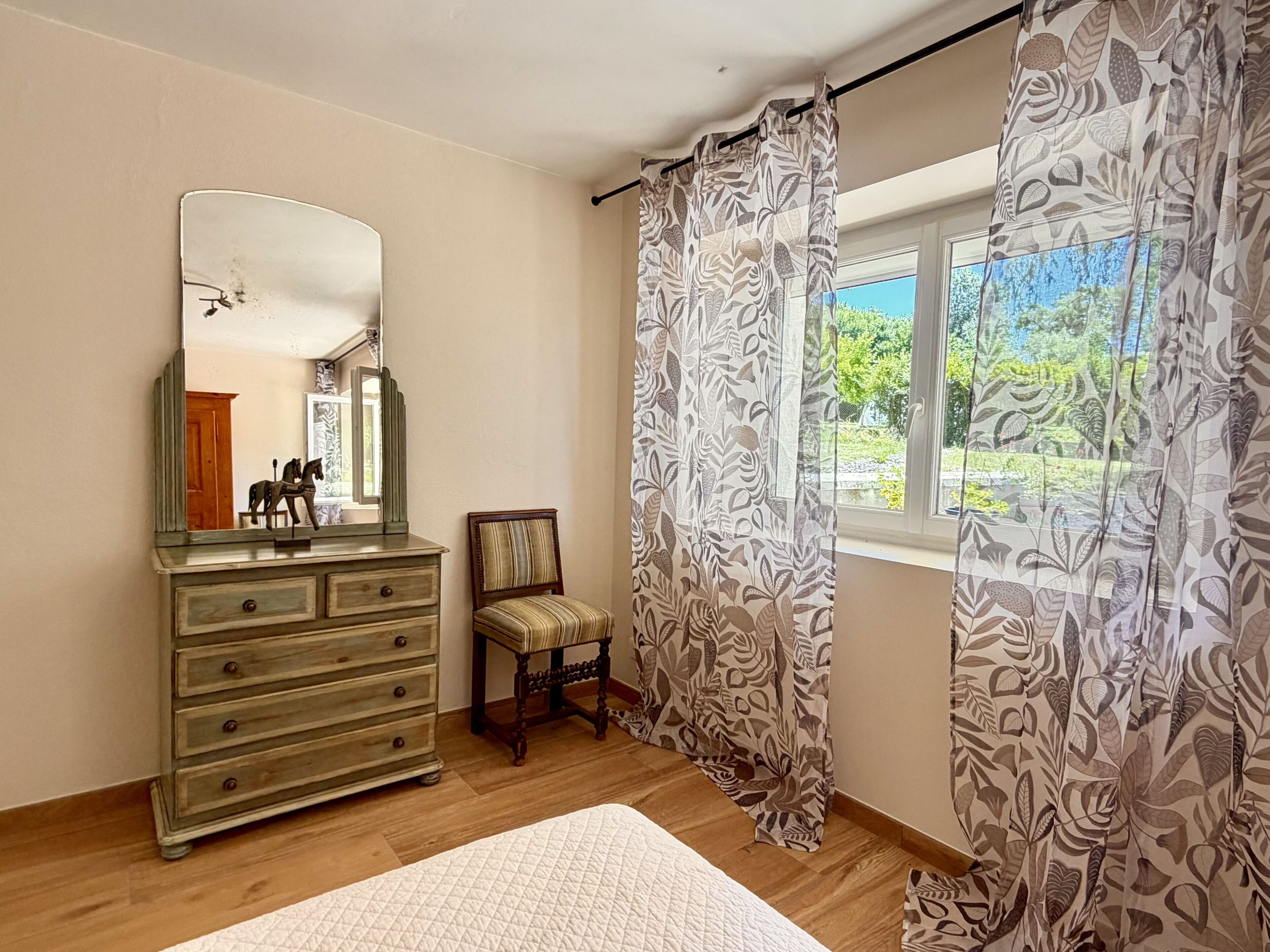 Un dormitorio con un aparador de madera, una silla, una cama con colcha, un espejo y una ventana con cortinas.