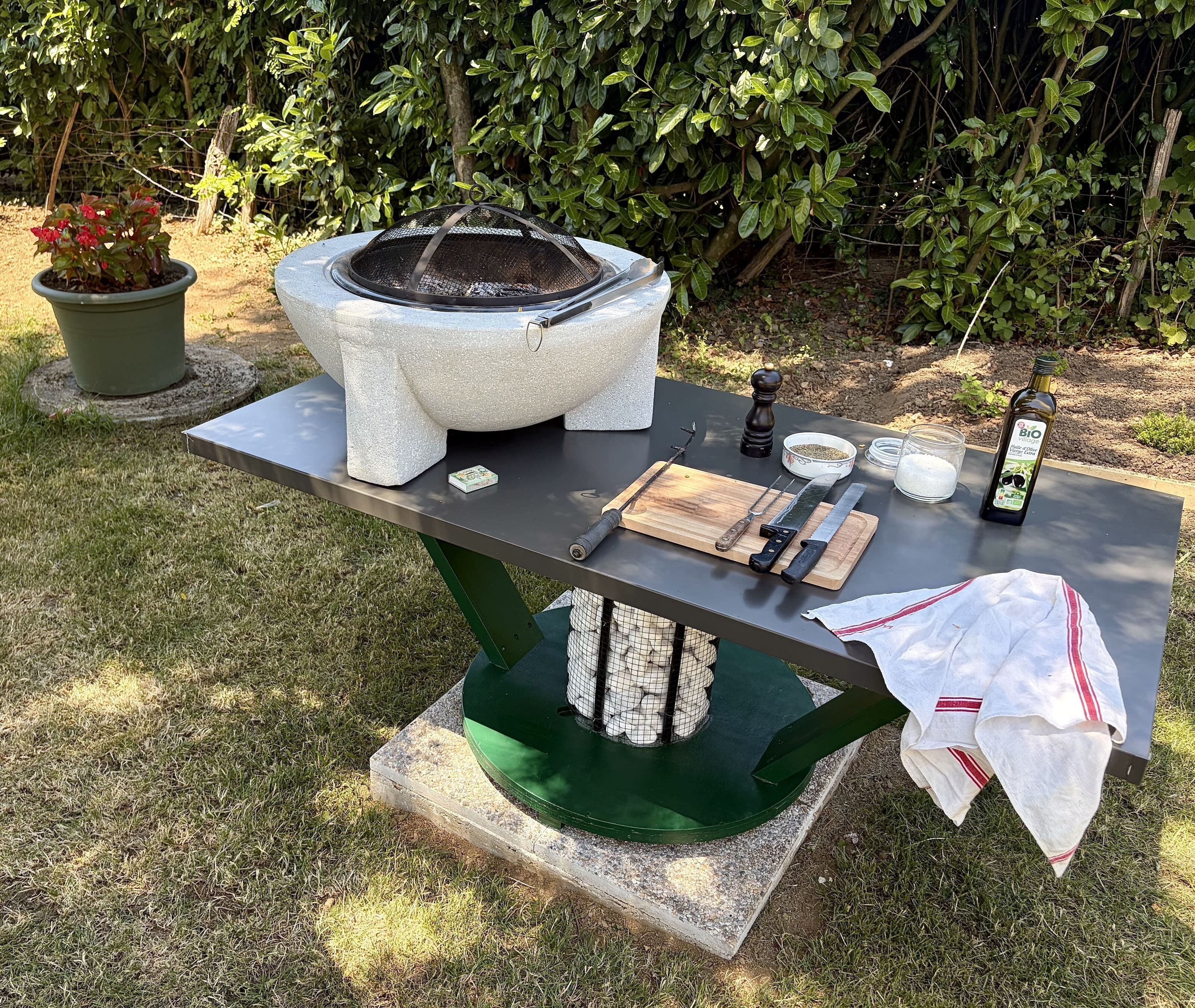 Een buitenkeuken met een vuurkorf van natuursteen, een tafel met kookgerei, een fles olijfolie en een bloempot.