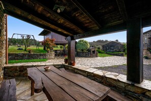 A rustic outdoor patio with a wooden table and benches, stone walls, and a view of a garden and buildings in the distance.