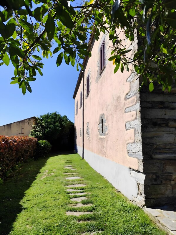 A narrow pathway leading to a building with a stone base and a pink upper wall, surrounded by greenery and a clear sky.