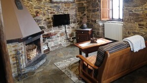 Habitació de parets de pedra amb una llar de foc, un televisor fixat a la paret, una taula de fusta, una cadira amb coixí i una finestra amb persianes de fusta.