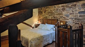 Un dormitori amb paret de pedra, bigues de fusta, un llit amb llençol de retalls, una tauleta de nit i una làmpada.