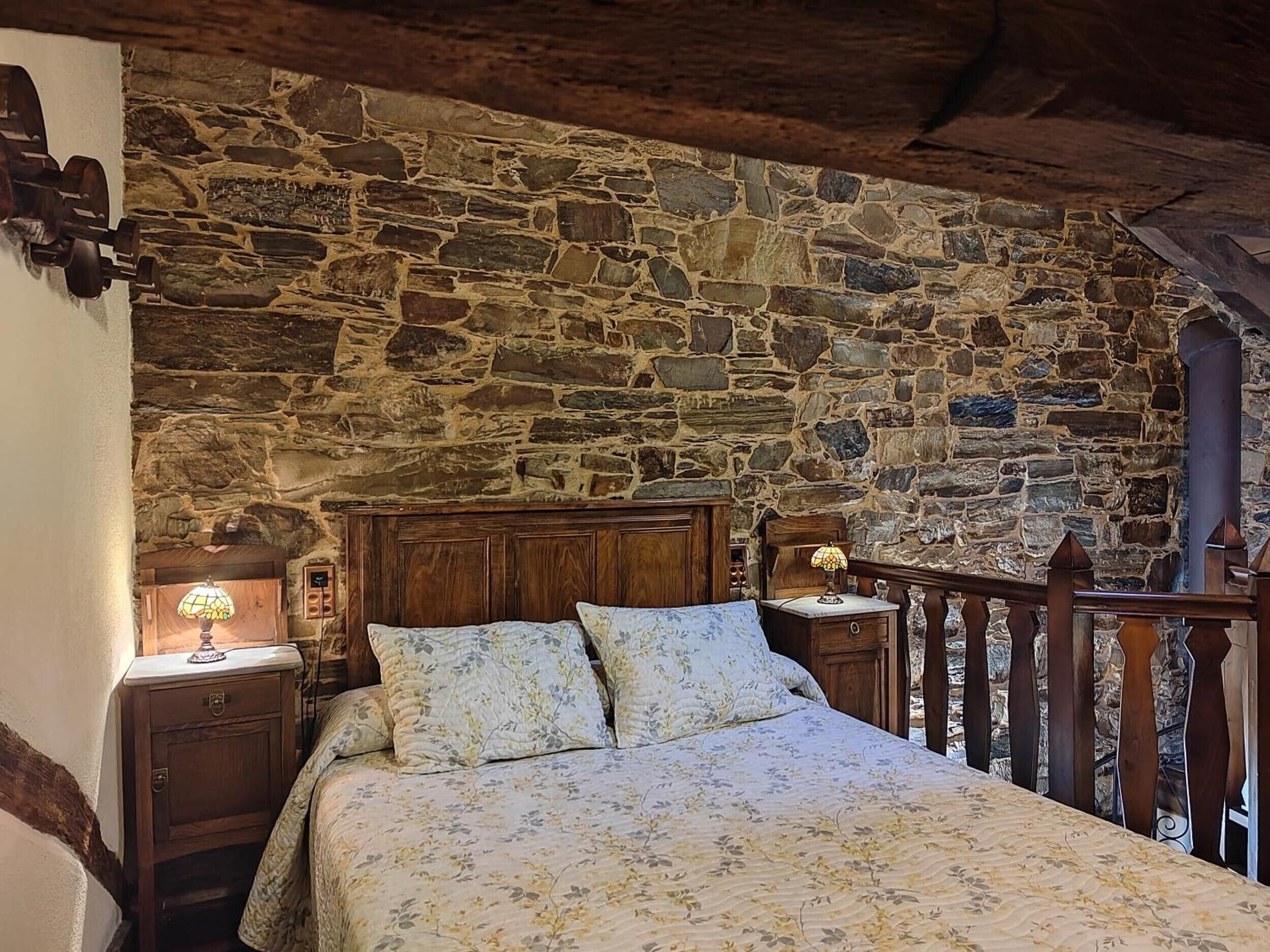 Une chambre avec un mur en pierre, une tête de lit en bois, des tables de chevet avec des lampes et un lit avec une literie à motifs floraux.